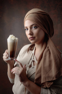 年轻美丽的女人在棕色头巾拿起一杯咖啡与奶油。完美的妆, 褐色的背景。型号加尺寸