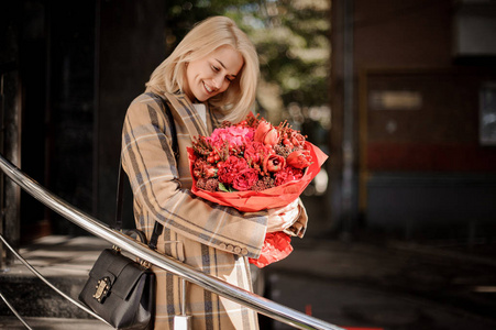 快乐微笑的金发女郎穿着格子大衣, 秋天的时候有一大束红色的花朵。