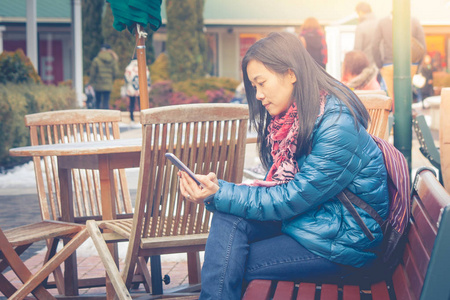 旅游寒假理念 亚洲女子旅行者坐在木制长板凳外, 玩她的智能手机