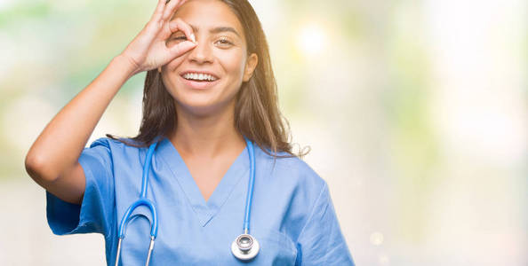 年轻的阿拉伯医生外科医生妇女在孤立的背景做 ok 手势与手微笑, 眼睛看通过手指与幸福的脸