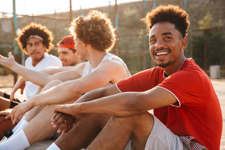一群年轻积极的多民族男子篮球运动员休息在运动场上, 谈论