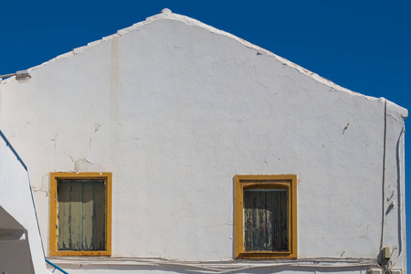 建筑与明亮的白色旧立面。两个窗户, 黄色的框架和木板, 而不是玻璃。屋顶的三角形。背景明亮的蓝天。希腊克里特的阿基欧斯尼古拉斯