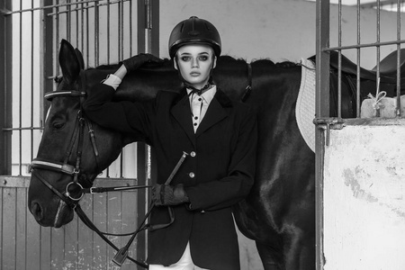 穿着赛马装备的漂亮女子在马厩运动和时尚概念上摆在大黑马附近