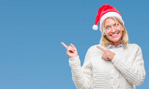 中年金发女子戴着圣诞节帽子在孤立的背景微笑和看着相机指着两个手和手指到一边