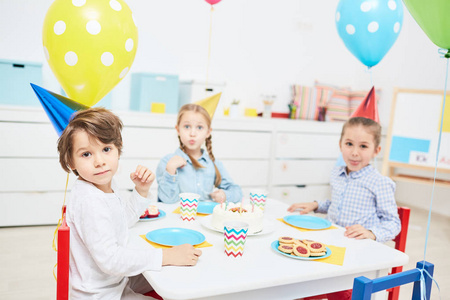 一群可爱的孩子在生日帽子坐在节日桌上与饮料和曲奇饼在幼稚园