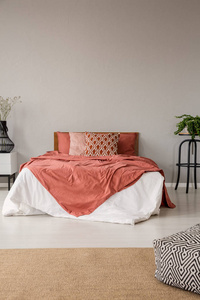 脚凳在地毯的最小灰色卧室内饰与红色床与枕头之间的植物。真实照片