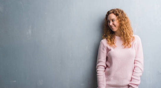 年轻的红发女人穿上灰色的垃圾墙, 穿着粉红色的毛衣, 脸上带着微笑, 表情自然。笑的自信
