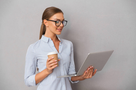 快乐的年轻女商人站在灰色背景, 工作在笔记本电脑, 喝咖啡