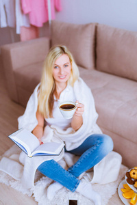 美丽的微笑金发女子坐在沙发旁边, 在右手和杯子咖啡在家里的左手保持一本书