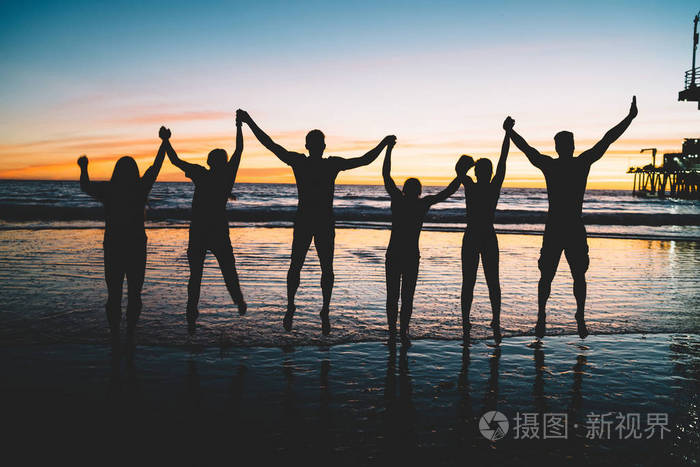 快乐的年轻男女在日落时分 牵着对方的手站在沙滩上 剪影一群人一起