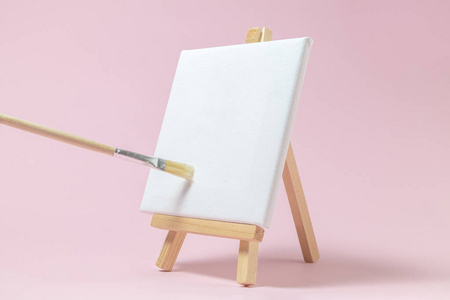 空白艺术板帆布与木立场微型和画笔在纯粉红色背景。艺术设备简约理念