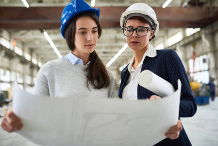 肖像两名女工程师穿着 hardhats 在现代工厂车间举行平面布置图