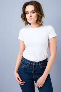 穿着白色 t恤衫和高腰蓝色牛仔裤的时尚年轻女孩的肖像在演播室灰色背景下摆姿势