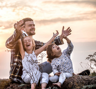 幸福美满的家庭, 爸爸和两个小可爱的女儿在日落时坐在一座高高的美丽的山上, 家庭关系和友谊的概念
