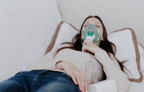 使用喷雾器和吸入器进行治疗。在沙发上躺着吸入口罩的年轻女子。前视图