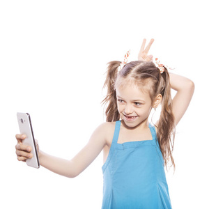 年轻的七岁的黑发女孩在蓝色礼服上的白色孤立的背景。她在她的手机上做自拍, 她脸上的乐趣和微笑的情绪