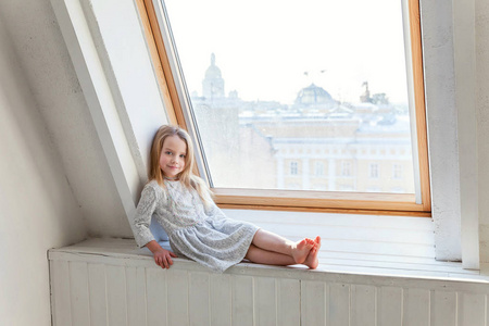 小可爱的可爱的微笑的女孩, 白色的礼服坐在窗台上, 在明亮的客厅在家里思考