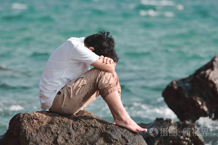忧郁的年轻亚裔男子抱着膝盖坐在海边的岩石上