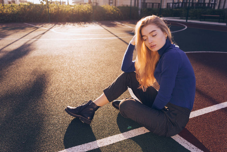 美丽的年轻长发金发女郎穿着蓝色毛衣坐在城市的运动场上, 摆姿势, 闭上眼睛。