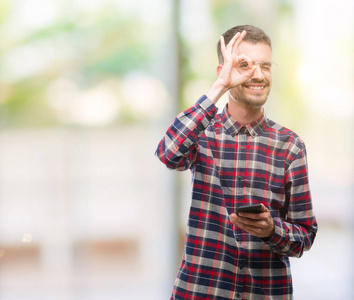 年轻的时髦成人男子持有智能手机与快乐的脸微笑做 ok 标志用手在眼睛看通过手指