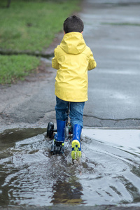 小男孩在雨衣和橡胶靴在踢滑板车, 后面看法