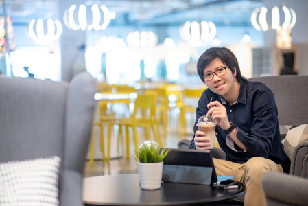 年轻的亚洲快乐商人穿着休闲风格喝咖啡与数字平板电脑在咖啡馆工作。自由生活方式或数字游牧概念