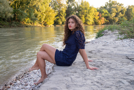在河岸上坐着长波浪褐色头发的女孩。细白沙。日落时温暖的太阳。那个女孩穿着一件蓝色的蕾丝连衣裙。