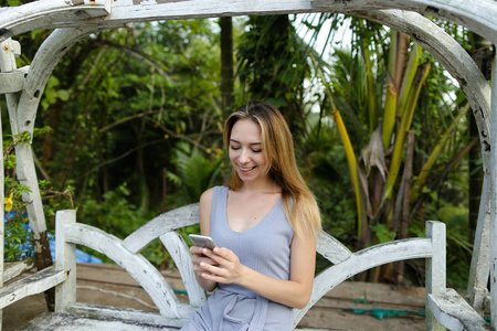 年轻女子骑秋千和使用智能手机在异国情调的花园, 棕榈在后台