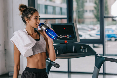 年轻的非洲裔美国女运动员喝水从运动瓶附近的跑步机在健身房