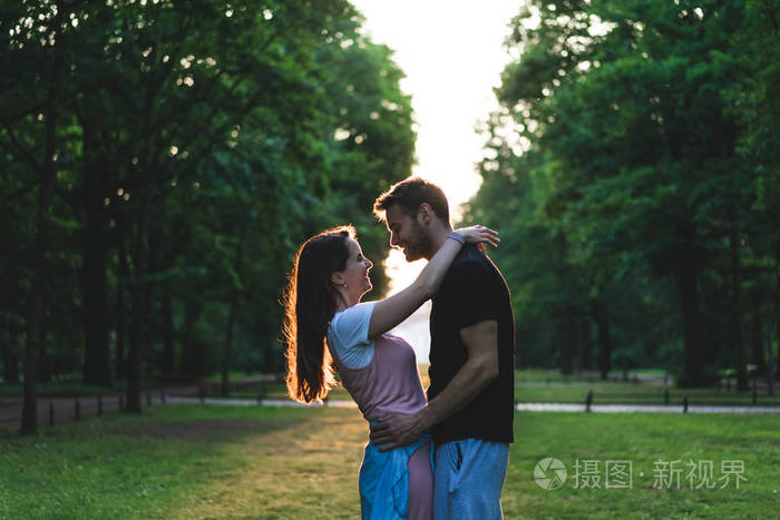 在公园的草地上拥抱彼此的微笑情侣的侧面视图