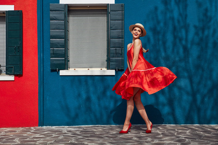 微笑的年轻女子在吹着风礼服, 高跟鞋, 草帽在头上, 在布拉诺岛附近的蓝色和红色的房子。墙上有树荫