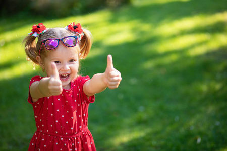 快乐滑稽的金发小女孩显示的目光竖起大拇指和良好的运气穿着鲜艳的红色礼服和太阳镜。复制, 空的文本空间