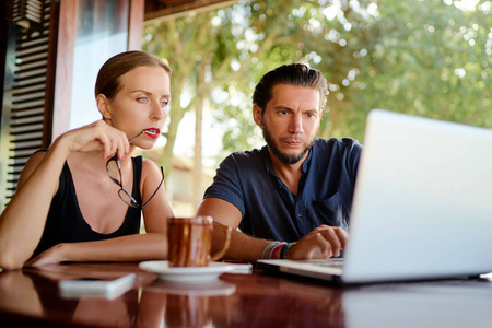 年轻的胡子男子和年轻女子一起工作在笔记本电脑上, 而坐在咖啡馆露台