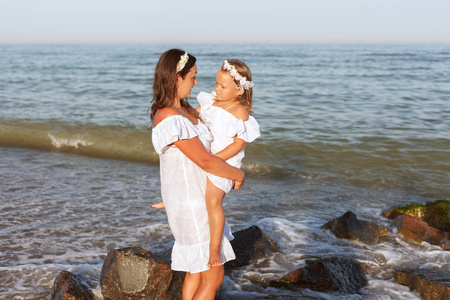 年轻的母亲和她的女儿穿着白色礼服站在沙滩上