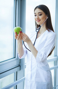 健康和美丽。医生营养师站在窗口附近的光办公室。女孩握着苹果的手和微笑。医生展示苹果