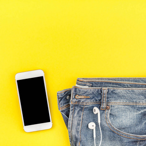 平躺的蓝色牛仔裤和智能手机耳机在大胆的黄色纸背景与复制空间。女性休闲服装的头顶景色。方形时髦看顶部视图