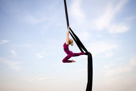 适合妇女在瘦瘦的衣服跳舞与空中丝绸在天空背景下, 体操运动员的空中丝绸训练