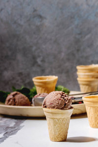 自制巧克力覆盆子冰淇淋在小华夫饼杯中, 用冷冻浆果, 新鲜薄荷, 碎黑巧克力和金属勺子在白色大理石厨房桌上的陶瓷板上