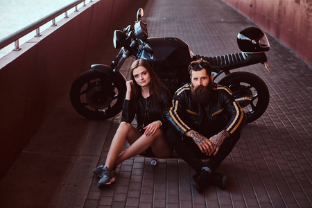野蛮的胡子车手在黑色的皮夹克和的黑发女孩坐在一起在一个滑板附近的定制复古摩托车人行道下桥