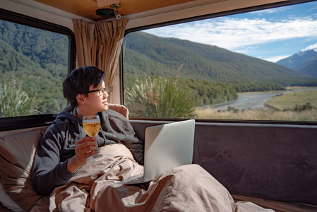 年轻的亚裔男子喝啤酒, 并与笔记本电脑在床上的露营车与山区风景景观通过窗口, 数字游牧概念