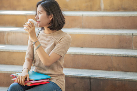 美丽的亚洲女大学生拿着她的书和一杯咖啡坐在楼梯上, 人们教育学习高中计划聪明的青少年概念