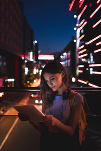 带耳机的年轻妇女的肖像在街道上用平板电脑与夜间城市灯光背景