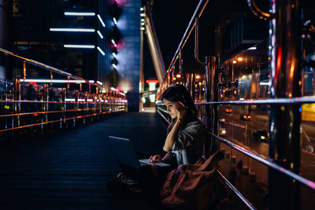 晚上在城市街道上使用笔记本电脑时, 女性在耳机中听音乐的侧面视图