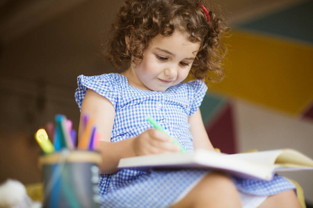 美丽的小女孩与深色卷曲的头发在蓝色的礼服朦胧画在着色书与毡尖笔在家里沙发上