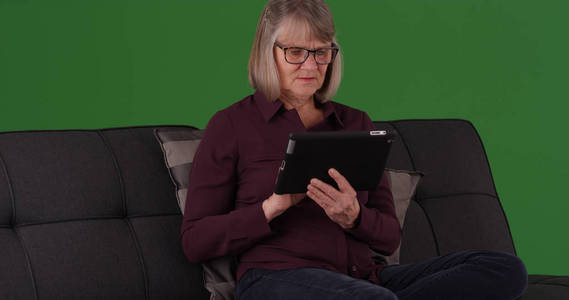 现代老年女性坐在沙发上使用平板电脑在绿色屏幕上