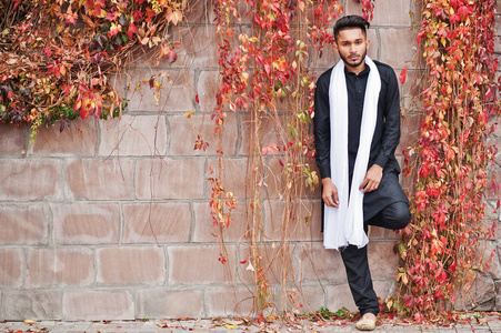 印度时尚男子在黑色的传统衣服与白色围巾摆在室外对红色叶子墙