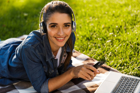 在公园的惊人的美丽的年轻女学生的形象使用笔记本电脑听音乐与耳机使用手机