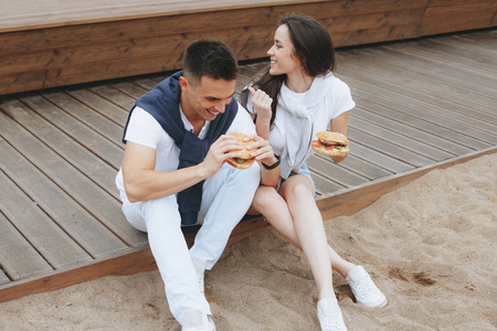 年轻美丽的积极夫妇坐在海滩上吃仓鼠。生活方式爱情和快餐