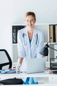快乐女医生在白色外套与听诊器在脖子站立在桌与膝上型电脑在办公室