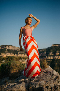 一个时髦的金发女孩在红色礼服摆姿势在古老的峡谷在日落。西班牙